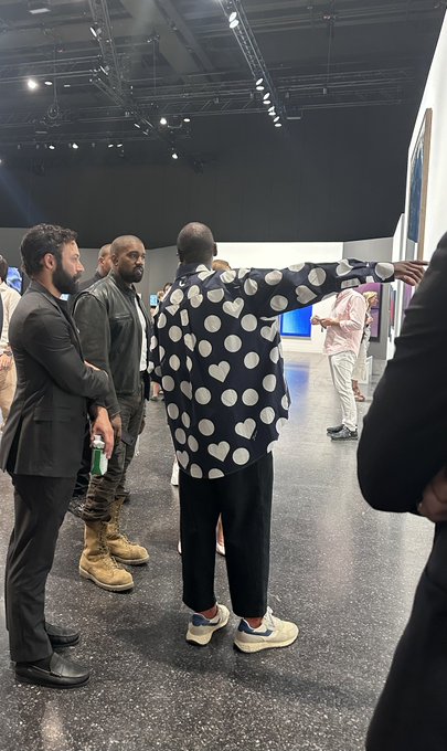 A man in a distinctive navy shirt covered in white polka dots and hearts stands with his arm extended at Kanye West's listening event, pointing toward another attendee in a gallery space.