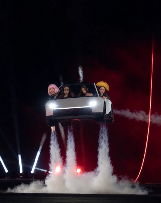 Four people wearing colorful wigs lean from a hovering white car during a smoky nighttime performance, with bright headlights, stage beams, and red lighting behind them.