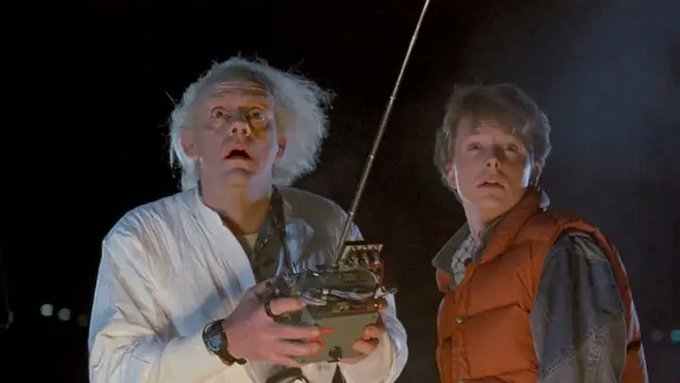 Two characters from Back to the Future, Doc Brown in a white lab coat and Marty McFly in an orange puffer vest, stare upward with alarmed expressions while Doc holds a remote-controlled device with an antenna.