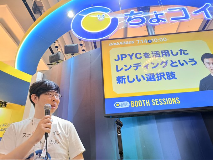 A man in glasses and a white t-shirt speaks into a microphone at a WebX 2026 booth session, with a screen behind him displaying 'JPYCを活用したレンディングという新しい選択肢' under a 'ちょコイ' sign.
