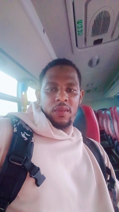 A man takes a selfie on a bus, with a backpack strap visible on his shoulder and empty red and black seats in the background, capturing a travel moment as he heads "Back to base."