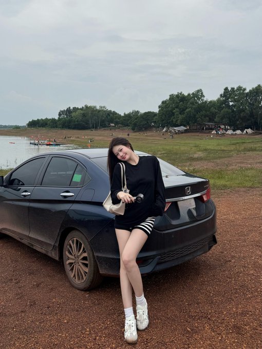 A young woman in a black outfit with striped shorts leans against a black Honda sedan parked on dirt near a lake, holding a small white purse and camera.