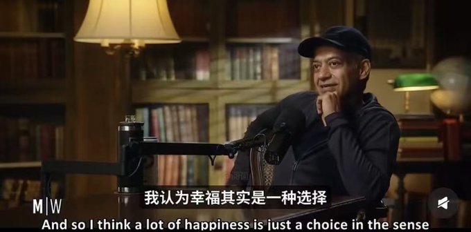 A man in a dark hoodie and cap speaks into a microphone in a room with bookshelves, with subtitles reading '我认为幸福其实是一种选择' and 'And so I think a lot of happiness is just a choice in the sense'.