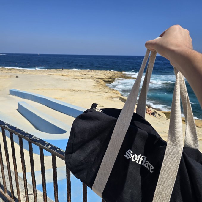 A hand holds a black "Solflare" tote bag against a sunny coastal backdrop featuring blue-painted concrete steps, sandy cliffs, and the ocean.