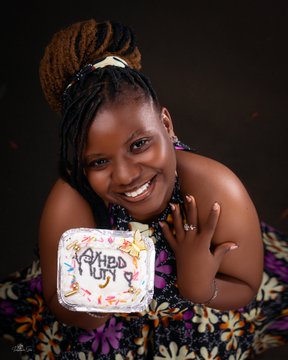 A smiling young woman with a stylish updo holds a small cake decorated with colorful flowers and the text "Happy Birthday" written in dark icing. The image captures a celebratory moment that aligns with the post, which highlights her special day alongside the Solana Stablecoin Summit in Uyo and a SuperteamNG trad ball event.