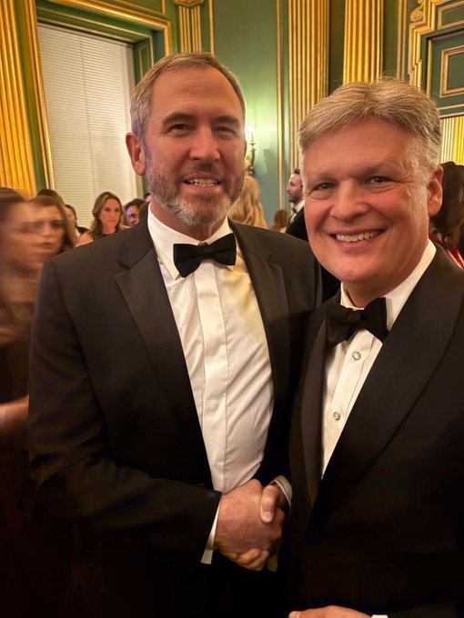 Two men in tuxedos smiling at a formal event with gold and green ornate decor in the background.