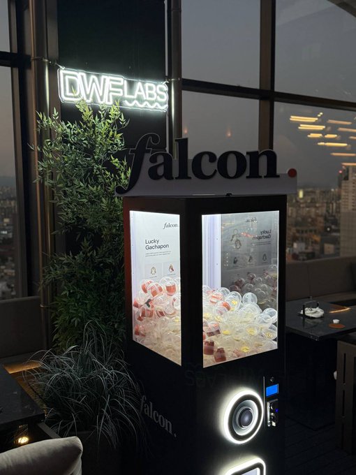 A 'falcon.' branded gachapon (capsule toy) vending machine with a 'Lucky Gachapon' display, situated indoors next to plants with a city view visible through large windows at night.