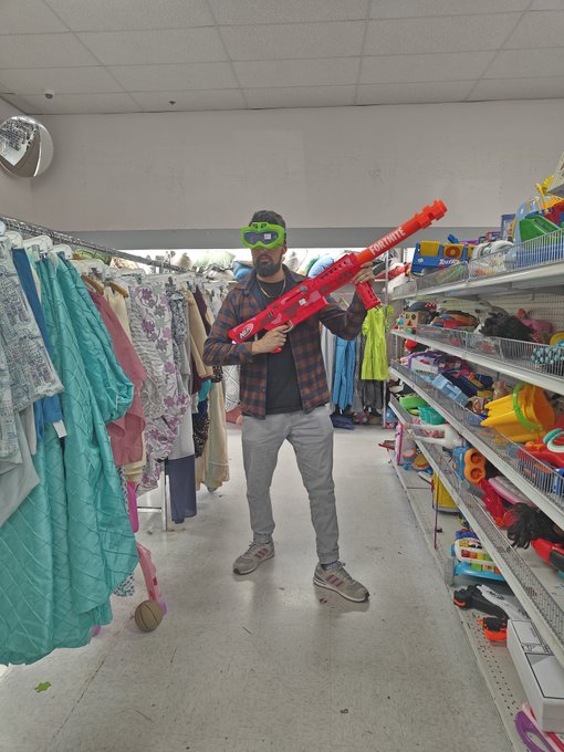 A man wearing green goggles stands in a thrift store aisle holding a large red Nerf Fortnite blaster, surrounded by clothing racks and shelves of toys.