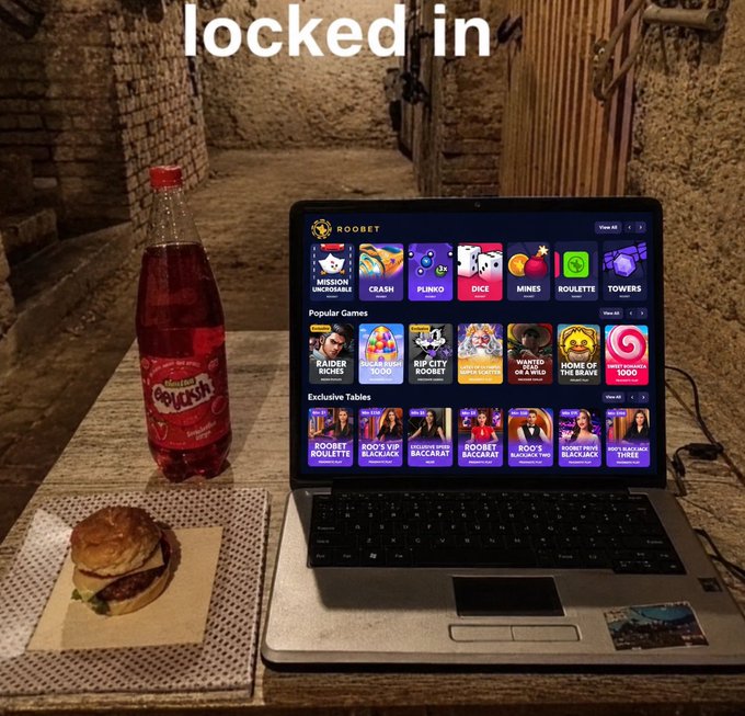A laptop displaying a Roobet gambling site with various casino games sits on a wooden table next to a bottle of red Fayrouz soda and a cheeseburger, with the text "locked in" overlaid at the top.