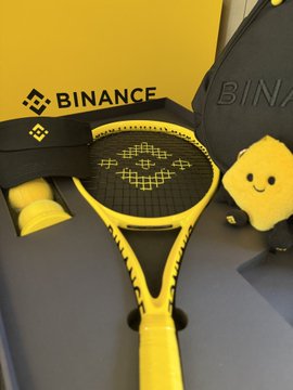 The image showcases a sleek yellow and black Binance-branded tennis racket with the iconic "BINANCE" logo and pixel art design on the strings, displayed alongside Binance merchandise. The accompanying post enthusiastically celebrates receiving this精美 (exquisitely crafted) Binance merchandise, suggesting the crypto exchange should open a周边 (merchandise) store accepting crypto payments.