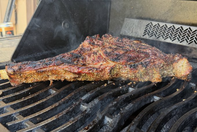 The image shows a large piece of cooked, medium rare steak placed on top of a black grill with smoke coming out from under it. The steak appears to be well-cooked and ready to be served. In the background, there are some buildings visible behind the grill, suggesting that this might be taking place in an urban setting or near a residential area.