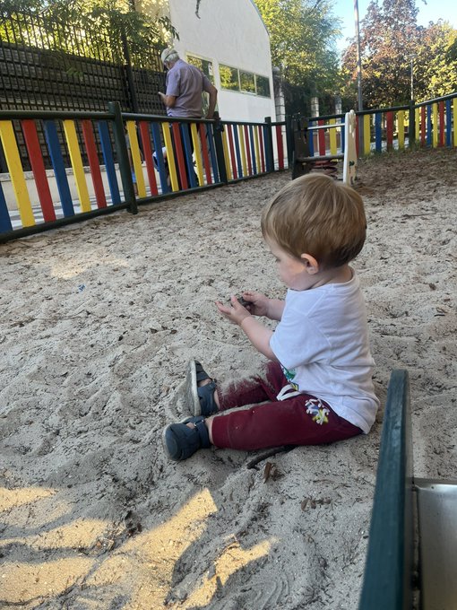 A young child in a white t-shirt and maroon pants sits in a sandy playground, playing with something in their hands, with an elderly person visible in the background.