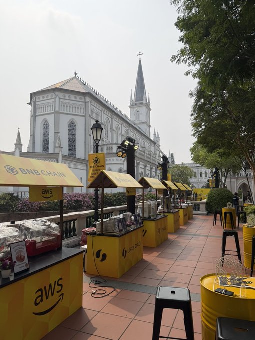 An outdoor setup featuring multiple yellow vendor stalls with 'aws' and 'BNB CHAIN' branding set up in front of a large white Gothic-style church building.