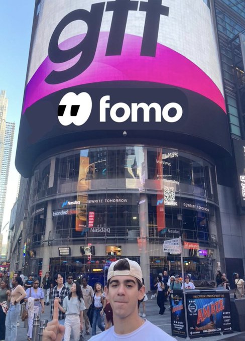 A young man in a white cap poses in front of the Nasdaq building in Times Square, where a massive billboard displays the "fomo" logo beneath "gtt" branding, while Nasdaq signs reading "Nasdaq" and "REWRITE TOMORROW" line the glass facade above the crowd.
