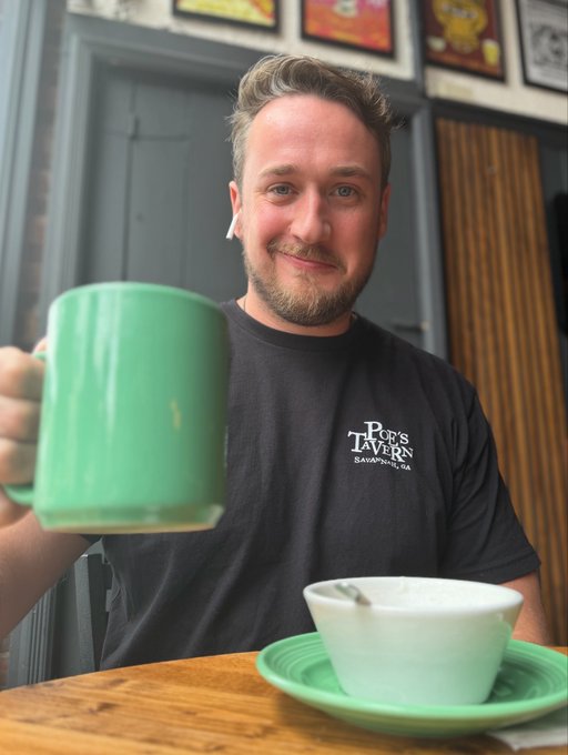 A smiling man with a beard and airpod wearing a 'Poe's Tavern Savannah, GA' t-shirt, holding up a large green coffee mug, with a matching green bowl on the table in front of him.