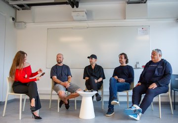 A diverse group of five panelists, including a man in a dark Flare Networks sweatshirt, sit on stage at the AI Connect event in Riga, Latvia, engaged in a discussion about AI development.