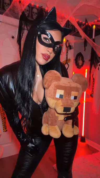 A woman in a black leather cat costume and mask holds a pixelated Minecraft-style brown bear plushie, with Halloween decorations and red lighting in the background.