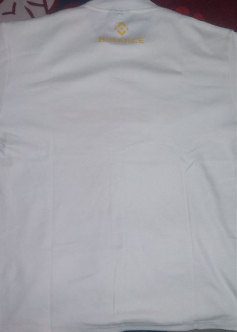 A white t-shirt with the gold 'BINANCE' logo is laid out on a surface.