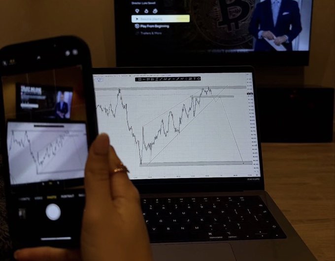 A hand holds a phone capturing a photo of a laptop displaying a Bitcoin price chart with a rising wedge pattern, while a TV in the background shows "Play From Beginning" next to a Bitcoin logo. The technical analysis setup reflects the post's observation that despite major positive catalysts like ETF approval, institutional adoption, and Trump's crypto advocacy, the market's wealth effect has diminished.