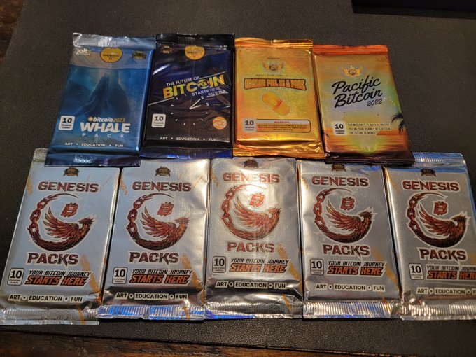 A collection of un-opened Bitcoin trading card packs displayed on a dark surface, including "WHAALE," "The Future of BITCOIN Starts Here," "10 SATOSHI," and "Pacific BITCOIN 2022" packs on top, with five silver "GENESIS PACKS" labeled "YOUR BITCOIN JOURNEY STARTS HERE" arranged in the bottom row. The image directly supports the post by showcasing the variety of collectible BTC trading card packs the collector owns.