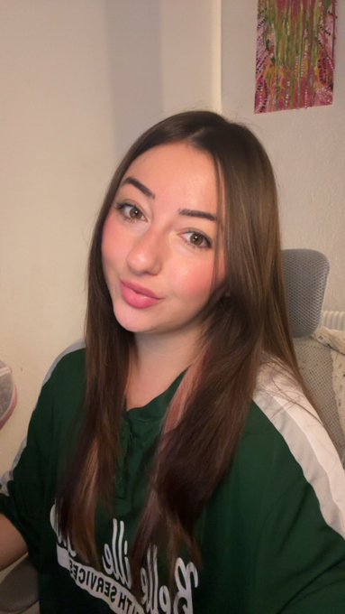 A woman with long brown hair featuring pink highlights smiles softly at the camera, wearing a green Hollister t-shirt while taking a selfie indoors.