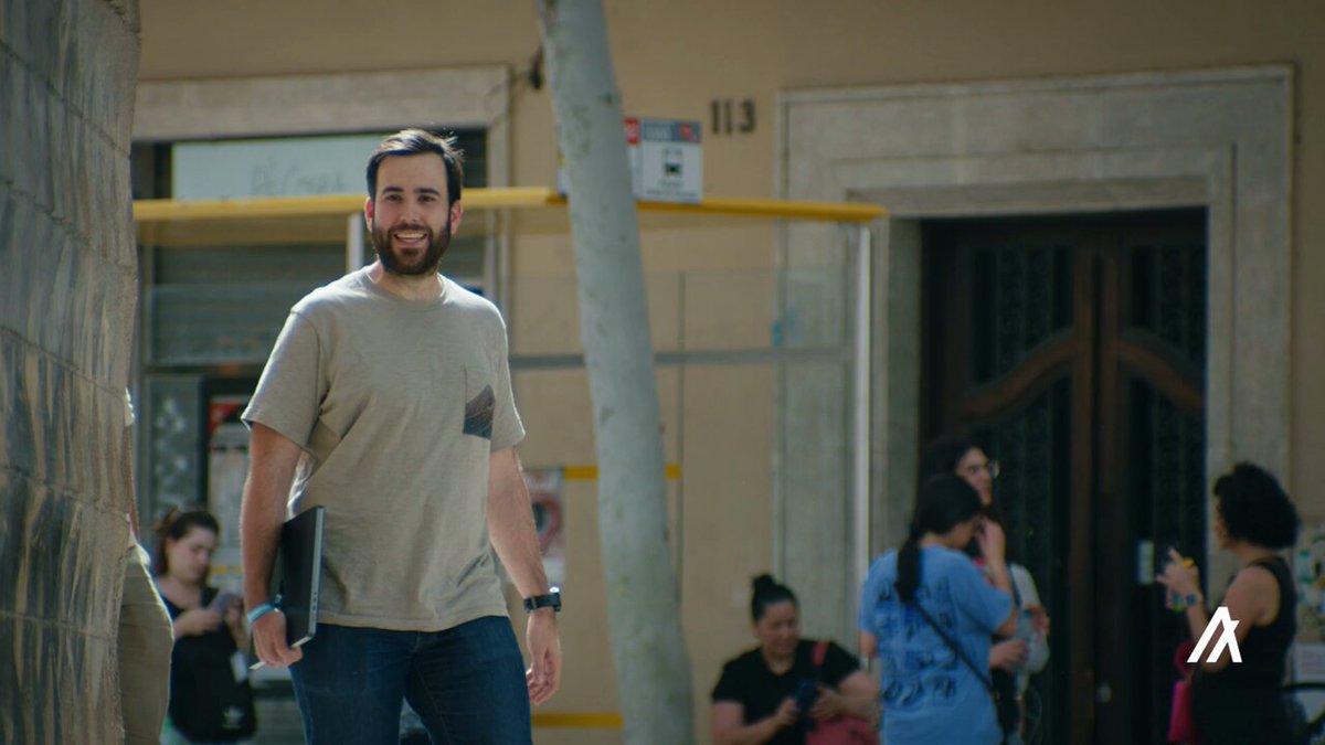 The image shows a man walking down a street with his laptop bag while smiling at the camera. The background includes other people going about their day, suggesting an urban setting. There are also signs for Alorand and Alorandbank, indicating that this scene takes place in a city where these organizations operate or have operations.