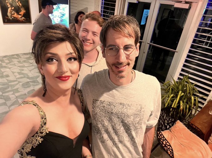 A selfie of three people at an indoor social event, featuring a woman with short dark hair in a black dress in the foreground, a smiling young man behind her, and a man with glasses on the right.