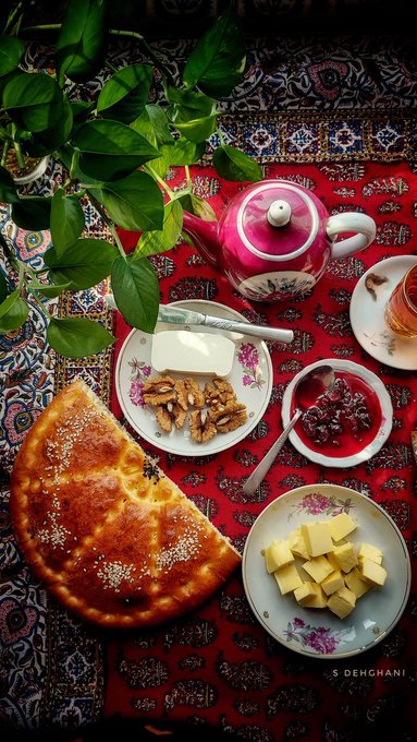 A flat lay photograph shows a Persian-style spread on a red patterned rug, featuring flatbread, butter, walnuts, jam, butter cubes, a teapot, and tea.