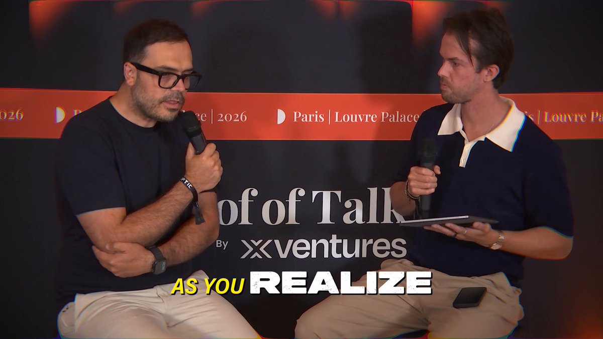 The image shows two men sitting next to each other with microphones in front of them. They are both wearing glasses and appear to be engaged in a conversation or interview. The background features a banner that reads "As you realize, there was no money lost. And this is by design." This suggests that the image might have been taken during an event related to cryptocurrency or blockchain technology, possibly at a conference or panel discussion.