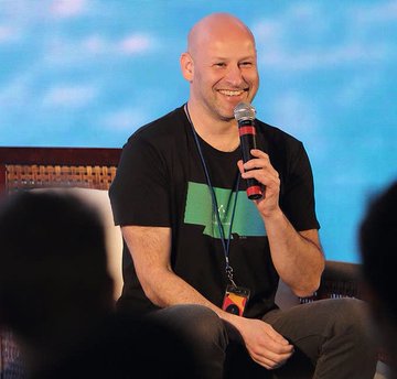 The image features a bald man wearing a black shirt with a green design sitting down while holding a microphone. He has an ID tag around his neck that reads "NEW". The background of the image shows a blue sky, indicating that he might be at an outdoor event or gathering. There are other people in the scene as well, suggesting that this is not just a one-person show but rather a larger event with multiple speakers and attendees.