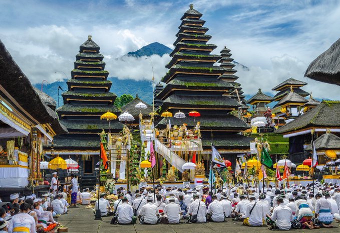 A large group of worshippers in traditional white attire gather at an ornate Balinese Hindu temple complex featuring multi-tiered pagoda-style towers (meru) adorned with yellow umbrellas and colorful flags, with a misty mountain in the background. The scene captures the spiritual essence of Bali's pervasive Hindu culture, where grand temple ceremonies bring communities together in elaborate displays of devotion.