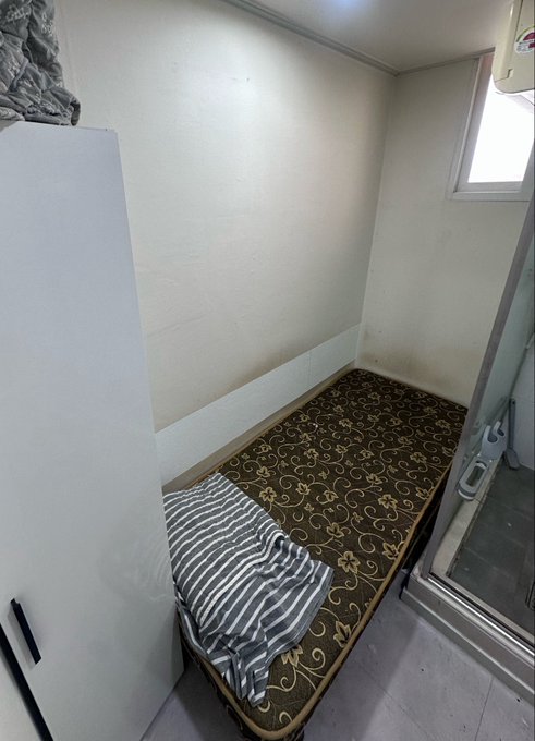 A small, narrow room containing a single mattress placed on the floor with a striped blanket, a small window, and white walls.