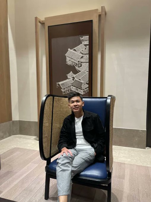 A man wearing a white t-shirt, black jacket, and grey sweatpants sitting on a tall blue and wood chair in front of a wall decoration depicting Asian architecture.