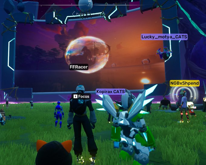 A group of players, including usernames "FFRacer," "Kopirax CATS," "Lucky__motya_CATS," and "NGBxShpend," gather in a vibrant OthersideMeta virtual landscape featuring a sunset over a floating planet with a "Focus" prompt visible. The scene captures the celebratory 1-year anniversary gathering on Apechain, showcasing diverse avatars mingling in the immersive digital world.