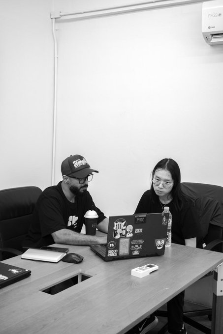 Two people, one wearing a cap and the other glasses, sit at a table working on a laptop covered in stickers, with a water bottle and coffee nearby, seemingly discussing Solana in what appears to be a casual office or studio setting.