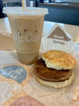 A McDonald's McCafé iced latte sits next to a Biscuit Sausage sandwich on a paper wrapper, with a triangular label reading "Biscuit Sausage" clearly visible. The caption references how the sweet iced latte pairs with the savory breakfast biscuit sandwich.
