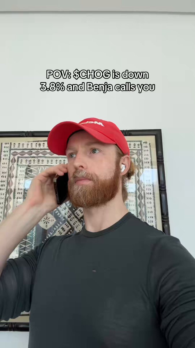 The image shows a man wearing a red baseball cap, talking on his cell phone while standing next to a picture frame. The caption of the image suggests that the man is sharing a funny moment with others.