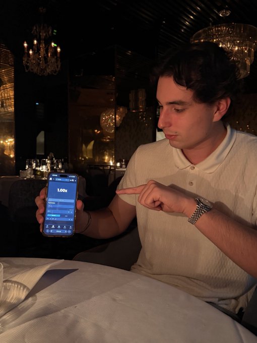 A man in a white polo shirt at an upscale restaurant displays his phone showing a Limbo betting app set at 1.00x, pointing at the screen as he solicits predictions on which multiplier to choose for a chance to win $100.