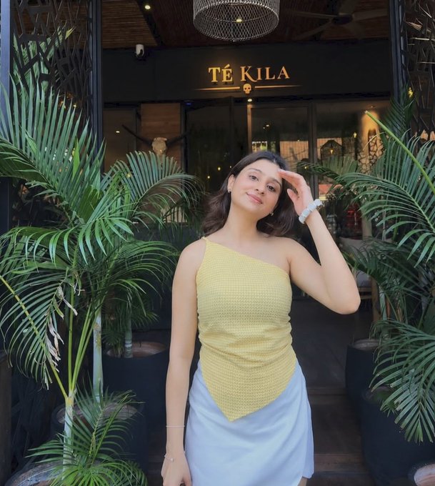 A woman wearing a yellow one-shoulder top and light blue skirt poses in front of a restaurant named 'TÉ KILA', surrounded by tropical plants.