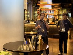 Two men stand in a dimly lit bar or lounge setting, one speaking while the other listens, with empty glasses on a small table in the foreground—capturing a casual yet engaged moment during the Variational-hosted dinner where the founder passionately discussed topics like TradFi liquidity and market maker mechanisms.