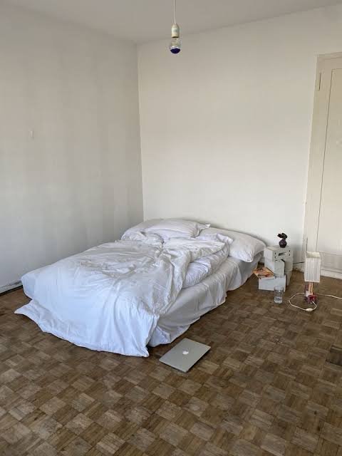 A minimalist bedroom with an unmade white bed, parquetry wood floor, a laptop on the floor, a small nightstand, and a bare pendant bulb hanging from the ceiling.