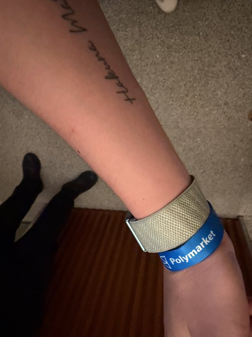 A person's forearm is shown wearing a blue "Polymarket" event wristband, with a partial "walk amongst" tattoo visible above it, while they stand on carpet near a wooden floor. The image captures the Polymarket event swag mentioned in the post, where the wearer is attending to collect perks for their group.