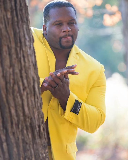 A man in a bright yellow blazer leans against a tree, hands clasped together in a gesture of grateful anticipation, mirroring the excitement of receiving a Reel Finance yield payout. The yellow color pops against the natural backdrop, emphasizing the "sweet" feeling of the 8% base APR in USDC, with a label on the jacket sleeve adding a touch of branded flair.