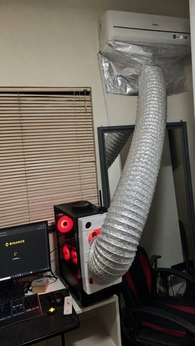 A homemade cooling rig channels air through a large flexible duct from a Haier wall-mounted air conditioner directly into a gaming PC with red fans, while the monitor displays the Binance logo and "Binance is working" — a humorous take on over-the-top PC cooling while crypto trading.