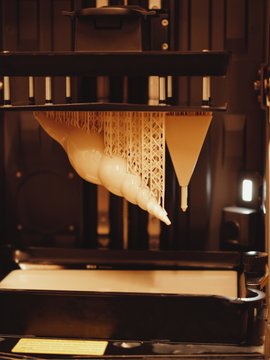 A close-up of a 3D printer in operation, showing a partially printed lattice-structured object being formed on the print bed under warm lighting.