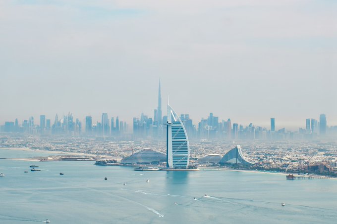 A hazy aerial view of Dubai's skyline featuring the Burj Al Arab and Burj Khalifa in the distance, with boats visible in the turquoise waters.
