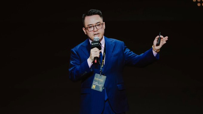 A man in a blue pinstripe suit and glasses speaks into a microphone on a dark stage, wearing a 'Speaker' lanyard and gesturing with his left hand while holding a clicker.