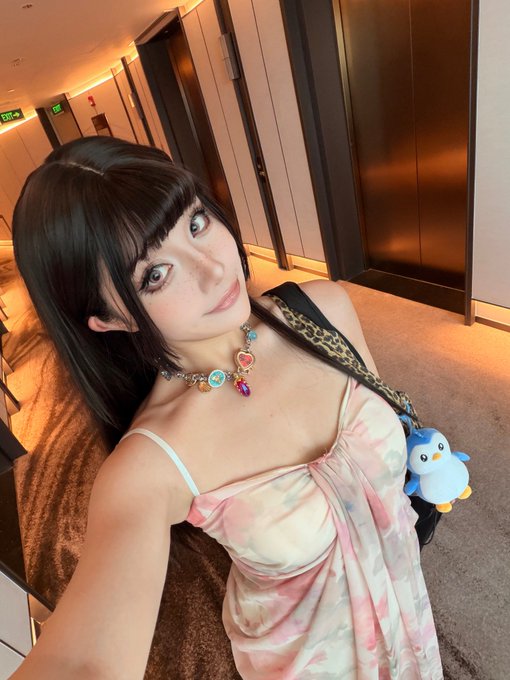 A selfie of a young woman with long dark hair wearing a floral pink dress and a colorful charm necklace, posing in what appears to be a hotel hallway with a small penguin plush accessory on her bag.