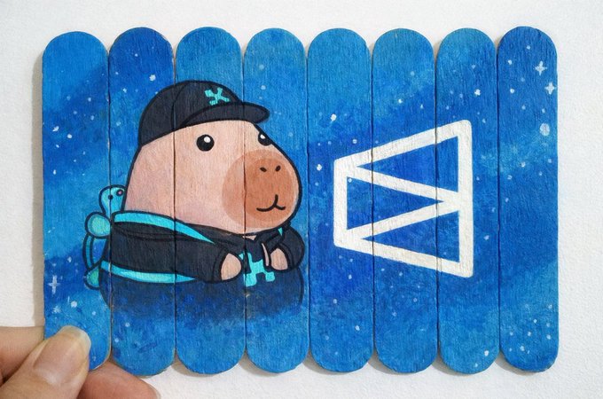 A hand holds a painted popsicle stick craft depicting a capybara wearing a cap and hoodie, next to a white emblem on a starry blue background.