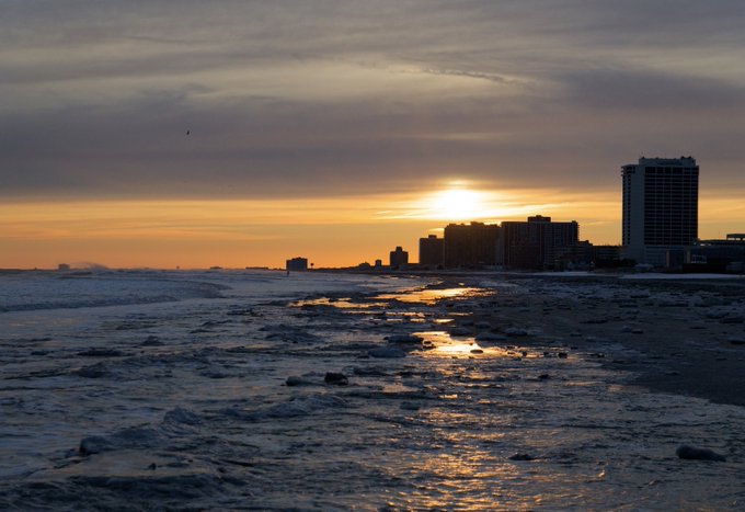 A breathtaking sunset over a coastal city, with the sun's golden reflection shimmering across the wet sand and ocean waves, while silhouetted high-rise buildings line the shore against a dramatic, cloud-streaked sky.